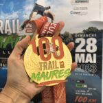 Médaille du 100 km du Trail des Maures dans une main avec en fond l'affiche du Trail des Maures