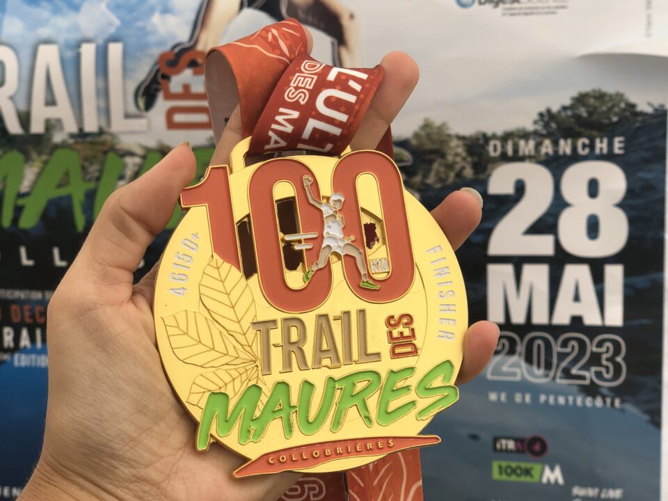 Médaille du 100 km du Trail des Maures dans une main avec en fond l'affiche du Trail des Maures