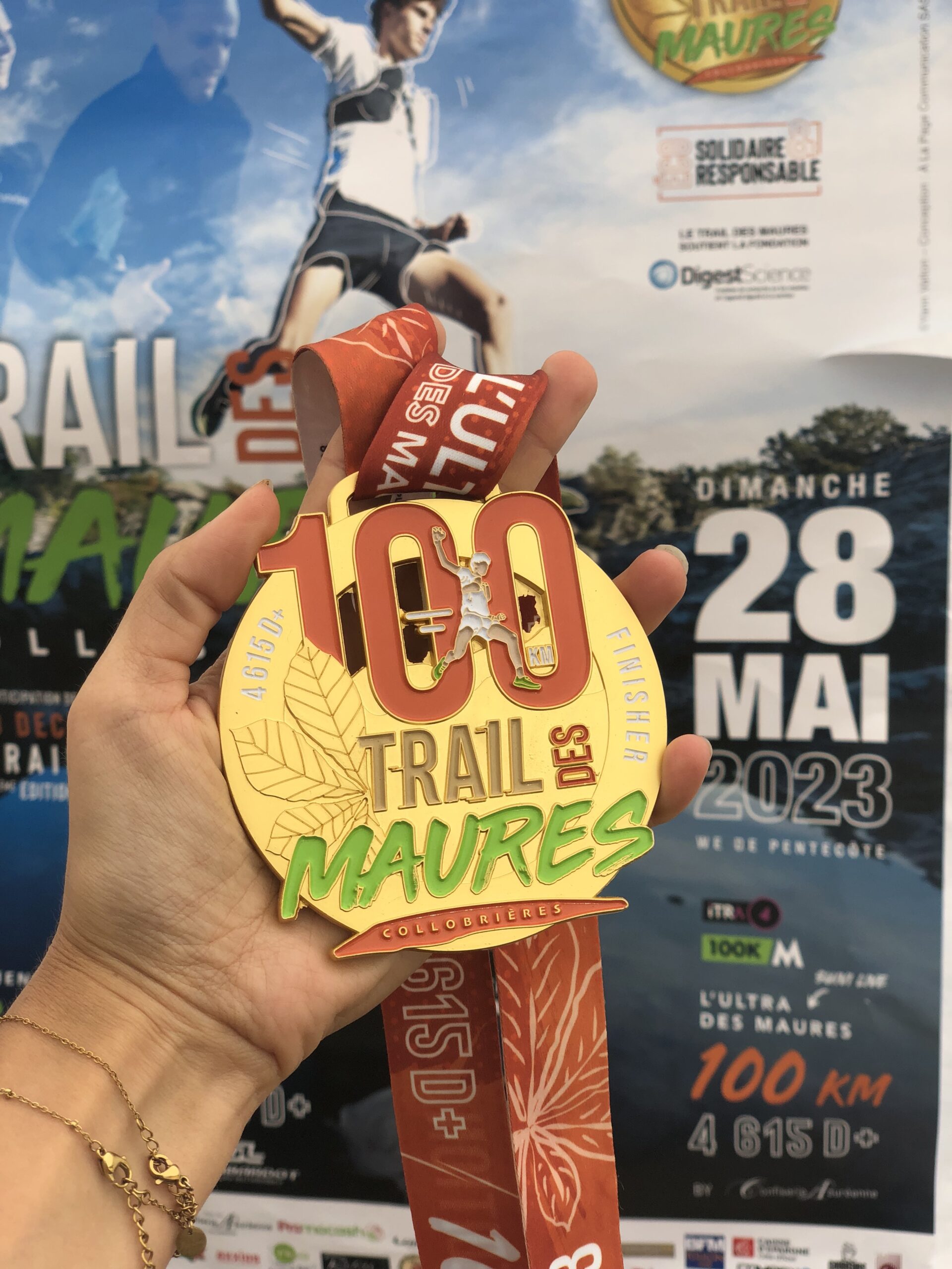 Médaille du 100 km du Trail des Maures dans une main avec en fond l'affiche du Trail des Maures