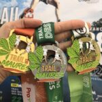 Trois médailles du Trail des Maures dans une main avec en fond l'affiche du Trail des Maures