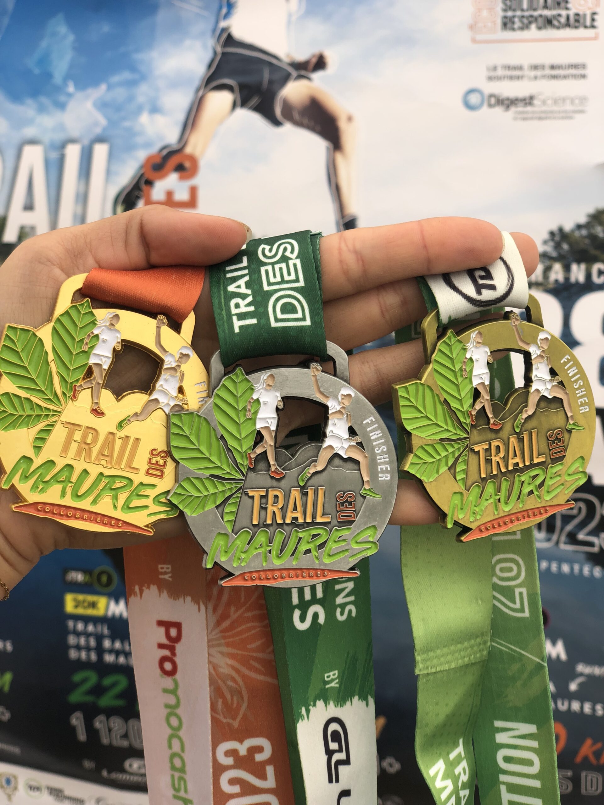 Trois médailles du Trail des Maures dans une main avec en fond l'affiche du Trail des Maures